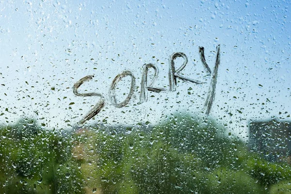 Word sorry on the window with raindrops. Background drop of water on the glass, blue sky, sun, city — Stock Photo, Image