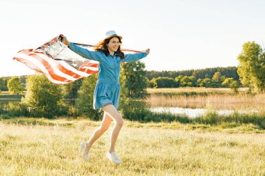 Kız Genç Amerikan bayrağı yaz yeşil çayırlarda günbatımında çalışan ile. Doğa arka plan, kırsal manzara