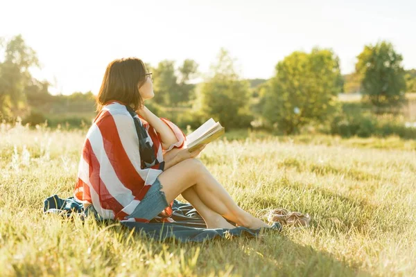 La mujer madura lee un libro, se apoya en la naturaleza. Sobre los hombros bandera americana, puesta de sol de fondo, paisajes rústicos, prado verde —  Fotos de Stock