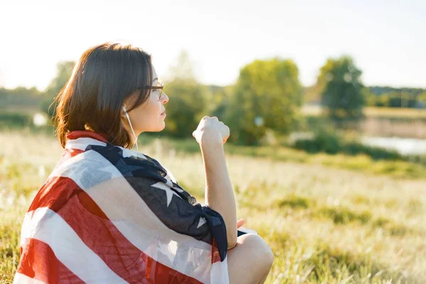 Mature woman listens to music, an audiobook on headphones, relaxes in nature. On the shoulders American flag, background sunset, rustic landscapes, green meadow — Stock Photo, Image