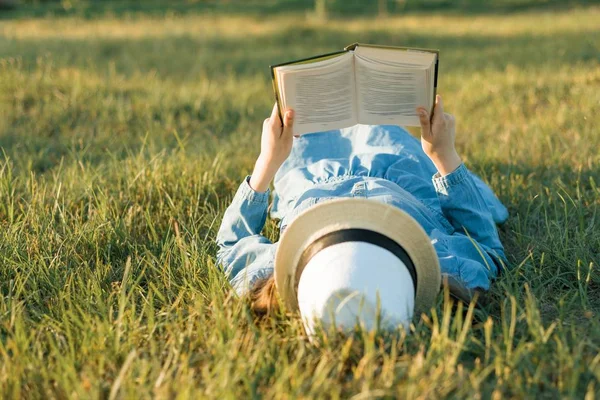 Dziewczyna w sukience i kapelusz znajduje się na zielonej trawie, czytanie książki. Od góry — Zdjęcie stockowe