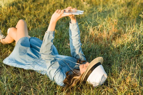 Retrato de adolescente de 14 anos deitado na grama. Menina de chapéu vestido, em seus fones de ouvido detém um smartphone, ouve música faz um photo.View selfi de cima — Fotografia de Stock