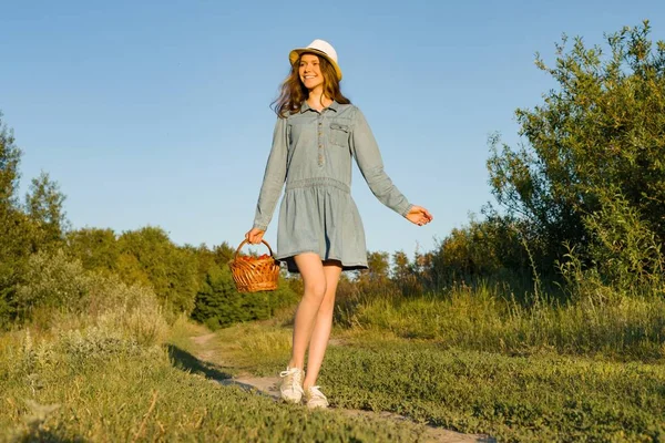 Venkovní letní portrét dospívající dívka v venkova venkovské silnici v šaty a klobouk pletený košík jahod. Přírodní pozadí, venkovské krajiny, zelená louka, stylu country. — Stock fotografie
