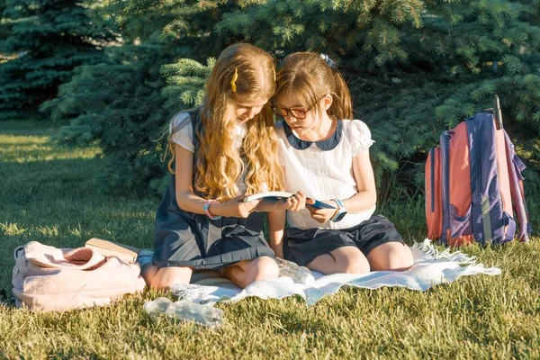 Dos amigas colegialas aprendiendo sentadas en un prado en el parque. Niños con mochilas, libros, cuadernos — Foto de Stock