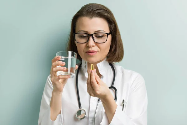 Portrait of female doctor with glass of water taking vitamin e capsule, fish oil, omega-3, pastel green mint wall background. Healthy lifestyle, nutritional supplements — Stock Photo, Image