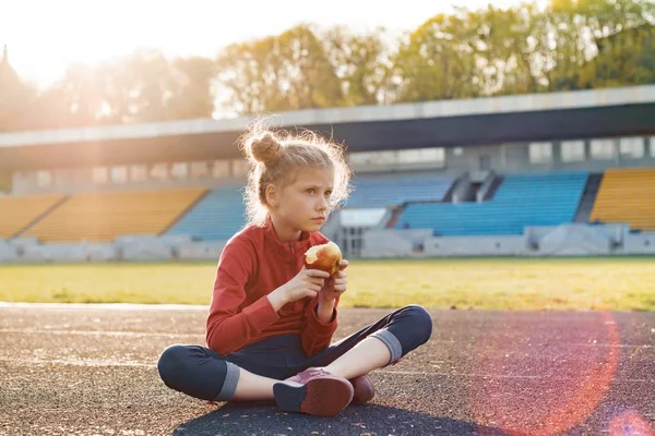 Egészséges életmód és az egészséges élelmiszerek fogalmát. Szép kislány gyermek sportruházat étkezési Alma ül a stadion, napsütéses tavaszi napon edzés után — Stock Fotó