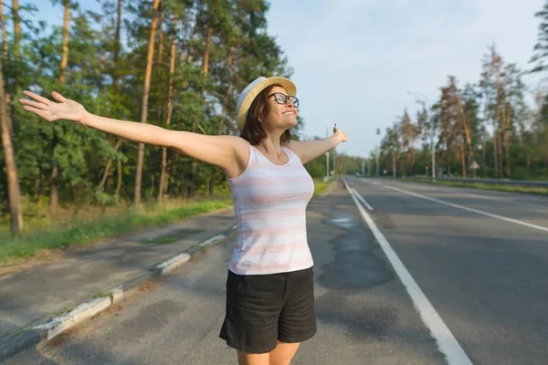 Smiling mature woman stands on the road, spread her arms out to the sides, enjoying freedom and sunset — Stock Photo, Image