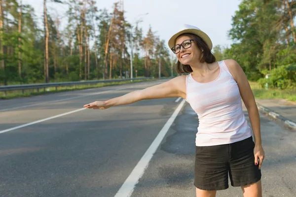 Outdoor portrait of smiling woman in hat standing on the road stopping a car, copy space — Stock Photo, Image