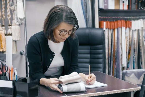 Workplace female designer, business woman in glasses with notebook pen and sample of fabric working in the office