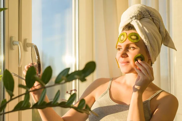Sorrindo jovem bela mulher fechar em casa perto da janela com máscara facial de frutas caseiro natural de kiwi no rosto, toalha na cabeça. Cuidados com a pele, cosméticos, cosmetologia, dermatologia . — Fotografia de Stock