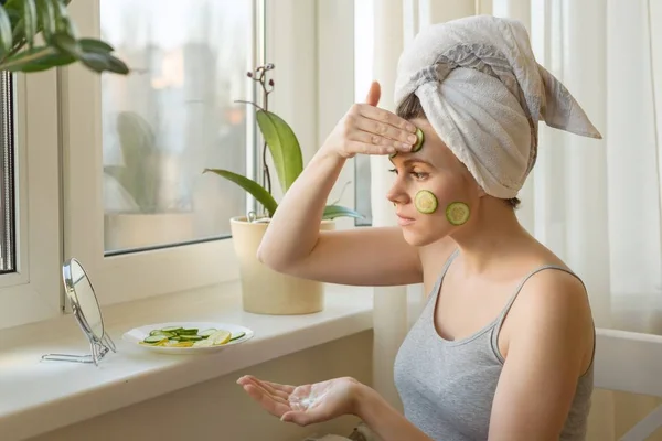 Ung kvinne i nærheten av vinduet som ser i speilet, bryr seg om ansikt med fløte og lager maske med agurk og sitron, håndkle på hodet. Hudpleie, kosmetikk, kosmetologi, dermatologi . – stockfoto