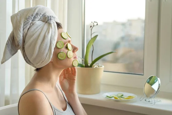 Jovem bela mulher close-up em casa perto da janela com máscara caseira natural de pepino no rosto, toalha na cabeça. Cuidados com a pele, cosméticos, cosmetologia, dermatologia . — Fotografia de Stock