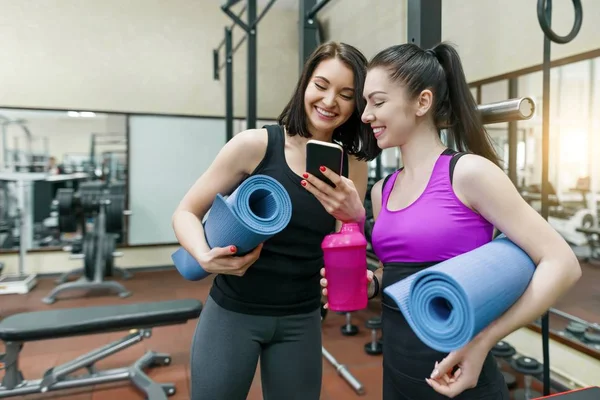Duas mulheres esportivas jovens no ginásio conversando sorrindo com tapetes de fitness e olhando para o telefone celular. Treinamento, trabalho em equipe, conceito de estilo de vida saudável . — Fotografia de Stock