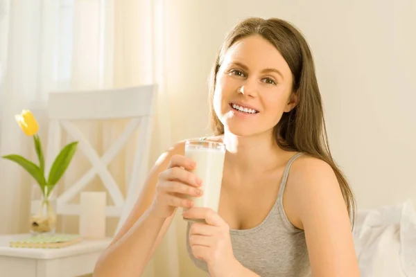 Junge schöne Frau sitzt zu Hause im Bett und trinkt Milch im Glas. — Stockfoto