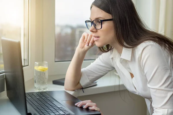 Joven mujer de negocios con gafas que trabajan en el ordenador portátil en casa, en el hotel, hablando en video, trabajando de forma remota . — Foto de Stock