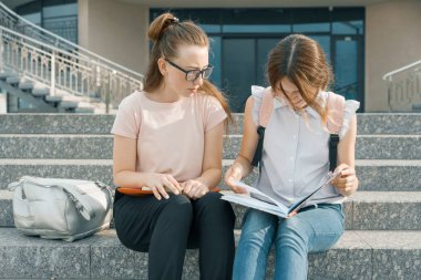 Sırt çantaları, kitap olan iki genç güzel kız öğrenciler açık portresi. Basamaklarında oturan, konuşurken, bir kitap haline arıyorum kızlar