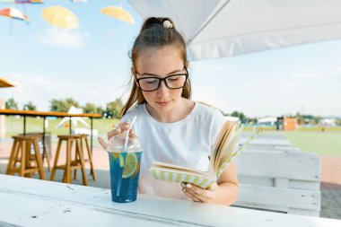 Kitap okuma ve yaz serinletici meyve kokteyli içerek kız yaz açık kafe, dinlenme genç gülümseyen genç kız.