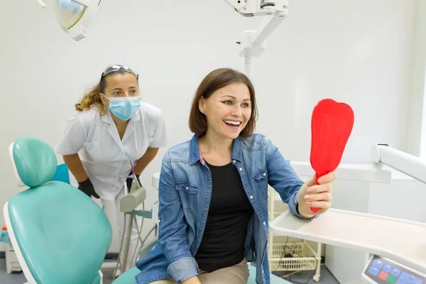 Mujer feliz paciente mirando en el espejo a los dientes, sentado en la silla dental . — Foto de Stock