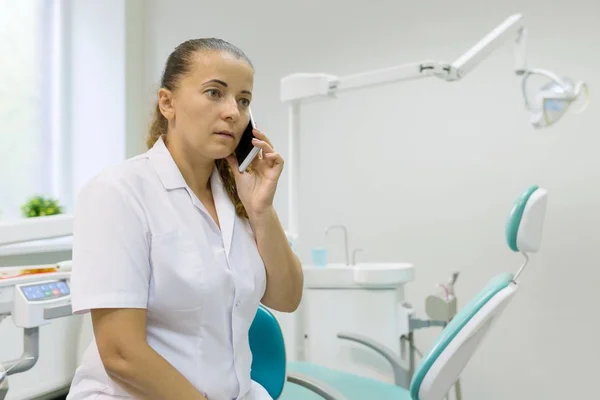 Portret van een vrouwelijke tandarts, arts praten op mobiele telefoon op tandheelkundige stoel achtergrond. Geneeskunde, tandheelkunde en healthcare concept — Stockfoto