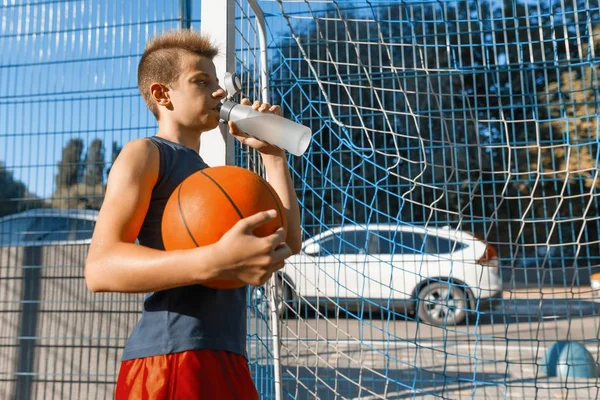 Adolescente niño jugando baloncesto callejero con pelota en la ciudad al aire libre cancha de baloncesto agua potable — Foto de Stock