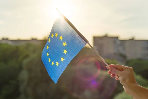 Ruka držící vlajka Evropské unie v otevřeném okně. Pozadí modré oblohy, silueta města, západ slunce — Stock fotografie