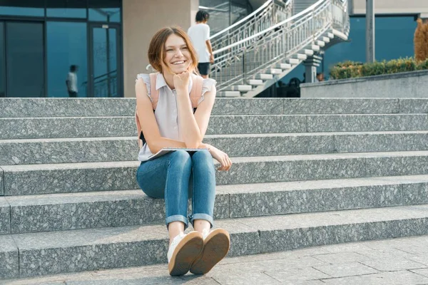 Retrato al aire libre de una joven y hermosa estudiante sonriente con mochila, sentada en los escalones de la ciudad —  Fotos de Stock