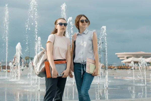 Retrato al aire libre de dos jóvenes hermosas estudiantes con mochilas, en la ciudad cerca de la fuente, las niñas mirando hacia adelante —  Fotos de Stock