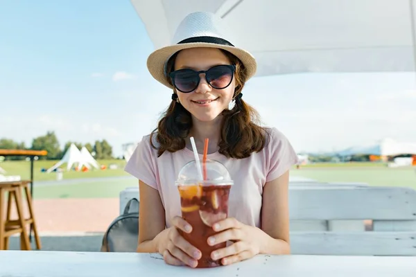 Joven adolescente con gafas de sol sombrero sonriendo y bebiendo cóctel de bayas fresco en un día caluroso de verano en la cafetería al aire libre — Foto de Stock