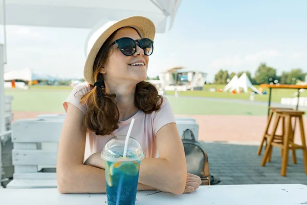 Joven adolescente en gafas de sol sombrero sonriendo y bebiendo cóctel de fruta fresca en un día caluroso de verano en la cafetería al aire libre —  Fotos de Stock