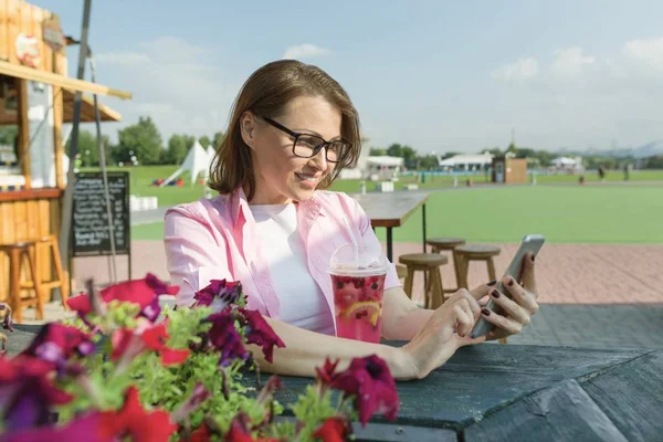Ώριμη γυναίκα αναπαύεται κάθεται στο τραπέζι στην καφετέρια του δρόμου το καλοκαίρι, στο κινητό τηλέφωνο, χαμογελώντας. Ζώνη φόντου πράσινο πάρκο ξεκούραση και ψυχαγωγία — Φωτογραφία Αρχείου