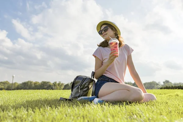 Chica joven en gafas de sol y sombrero bebidas bebida de bayas de verano con hielo sentado en la hierba verde, espacio para copiar, hora dorada — Foto de Stock