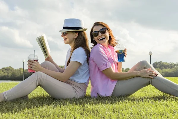 Общение родителей и подростков. Мать и дочь-подросток сидят на зеленой лужайке в парке и читают книгу, пьют прохладный коктейль и наслаждаются летним солнечным днем, золотым часом — стоковое фото