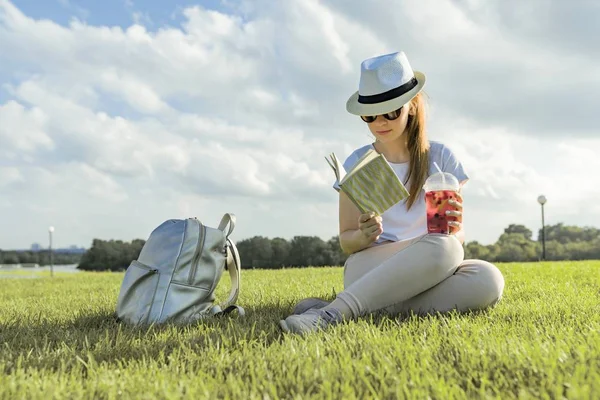 Jeune adolescente dans le chapeau et les lunettes lit le livre, boit boisson fraîche aux baies. Profite de l'été assis sur l'herbe verte dans le parc, heure d'or — Photo