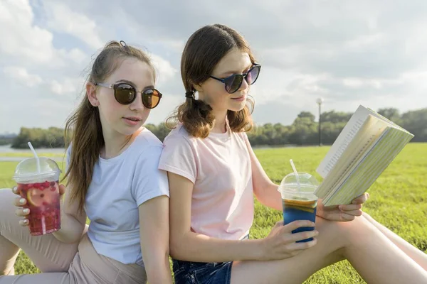 Día soleado de verano, dos novias adolescentes se sientan en el césped verde, beber cócteles, hablar, leer libro, reír, divertirse, hora de oro — Foto de Stock