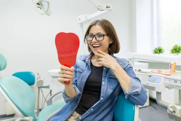 Щаслива жінка пацієнтка дивиться у дзеркало на зуби, сидячи в стоматологічному кріслі Ліцензійні Стокові Фото