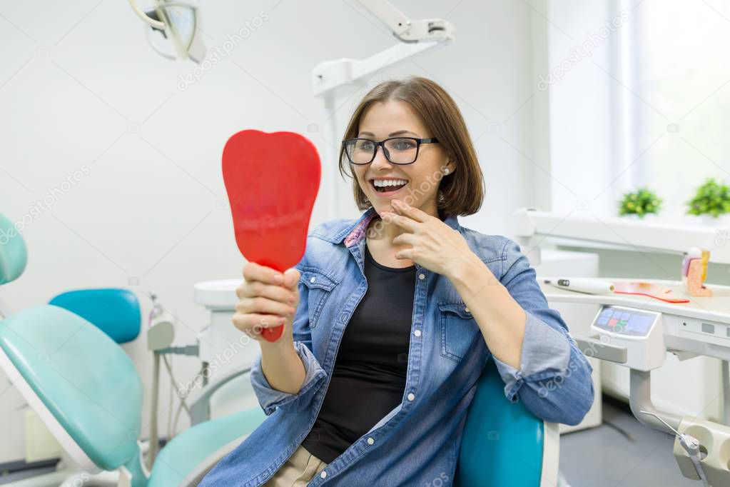 Happy woman patient looking in the mirror at the teeth, sitting in the dental chair