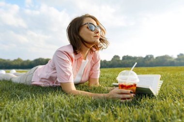 Olgun kadın yeşil çimenlerin üzerinde yatıyor. Kadın parkta yaz içeceği, kitap, dinlenme ve meditasyon, kopya alanı, altın saat