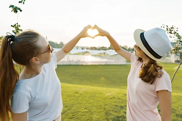 Twee meisjes vrienden Toon handen een hart symbool teken bij zonsondergang op een zomerdag in het park, het gouden uur — Stockfoto