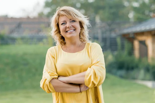 Außenporträt einer positiv selbstbewussten reifen Frau. Lächelnde Blondine in gelbem Kleid mit verschränkten Armen in der Nähe des Hauses — Stockfoto