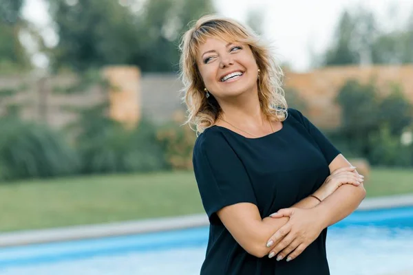 Retrato al aire libre de mujer madura segura positiva. Sonriente rubia con los brazos cruzados, fondo de piscina, residencia privada —  Fotos de Stock