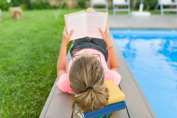 Adolescente de óculos lê um livro, piscina de fundo, gramado perto da casa. Escola, educação, conhecimento, adolescentes — Fotografia de Stock