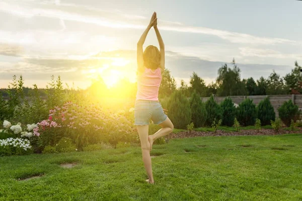 Ung tonårsflicka öva yoga, meditera över en sommar solnedgång bakgrund, på grönt gräs, gräsmatta nära huset. — Stockfoto