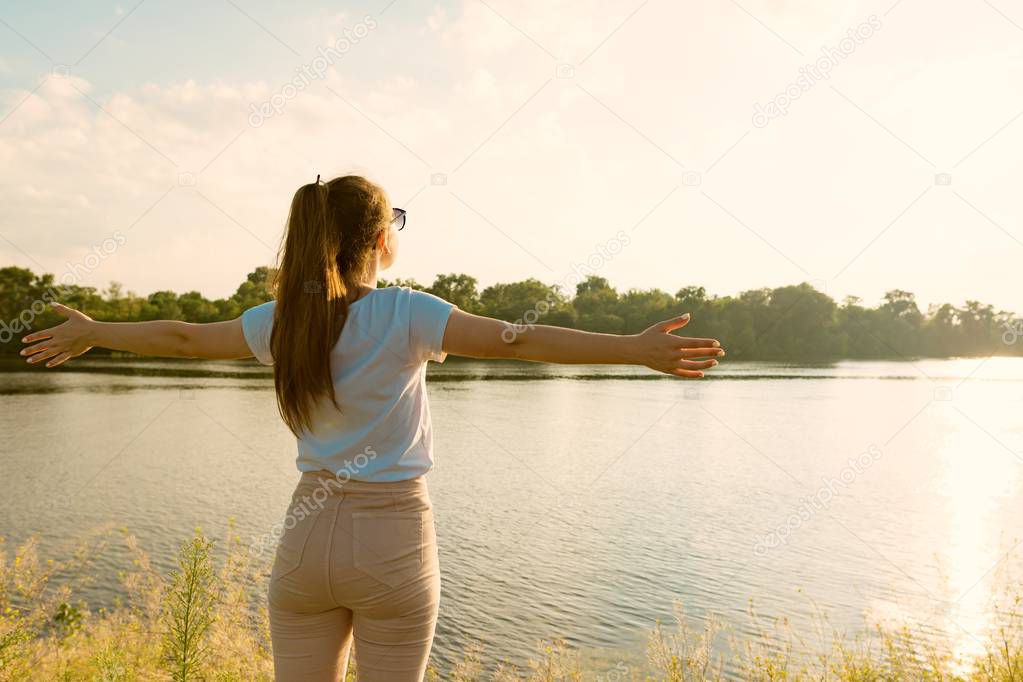Young girl enjoys the sunset on the river beautiful landscape, the back of a girl spread her arms to the sides, golden hour