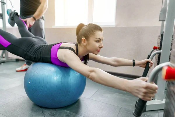 Young woman doing rehabilitation exercises with personal instructor using kinesi machine, fitness gym background. Kinesis technology, kinesitherapy, healthy lifestyle.