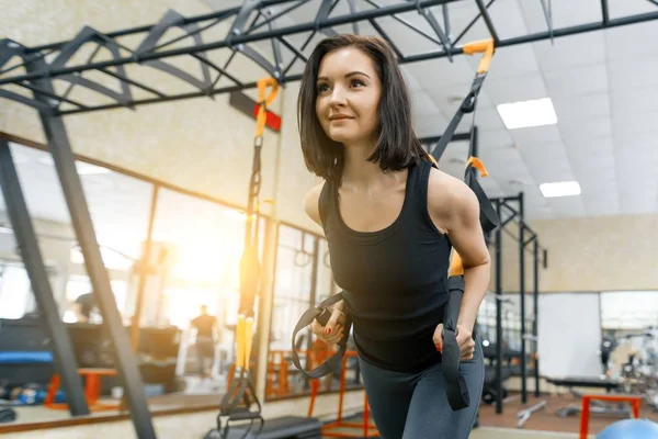 Young woman at the gym doing fitness exercises using sports straps system, holding hands by the loops. Fitness, sport, training, people concept.