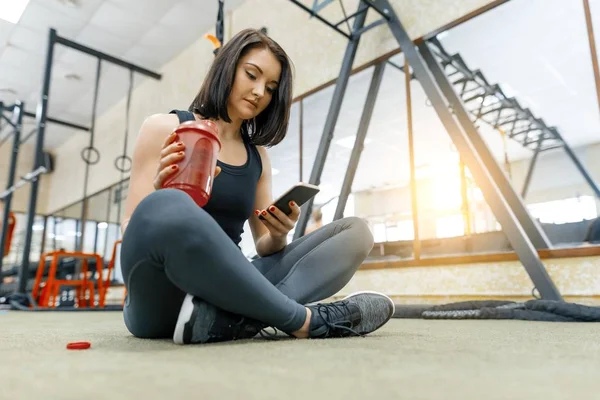 Junge Sportlerin, die sich nach den Übungen im Fitnessstudio auf dem Boden ausruht, Wasser trinkt und das Smartphone liest. Fitness, Sport, Training, Menschen, gesundes Lebensstilkonzept. — Stockfoto