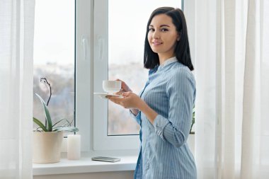 Pencerenin yakınında mavi gömlek kahve bir fincan ile genç güzel gülümseyen kadın portre