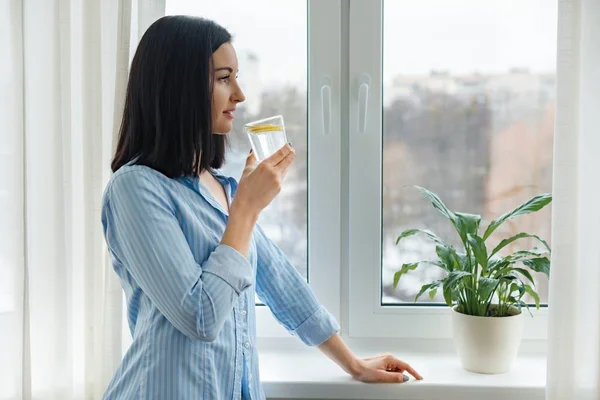 Jeune femme matin à la maison près de la fenêtre boire de l'eau avec du citron, boisson vitaminée en hiver printemps saison — Photo