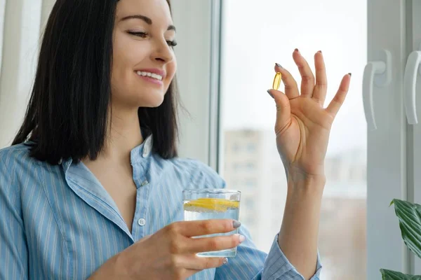 Mulher toma pílula com ômega-3 e segurando um copo de água doce com limão. Foto matinal da casa, perto da janela. Vitamina D, E, cápsulas de óleo de peixe. Nutrição, alimentação saudável, estilo de vida — Fotografia de Stock