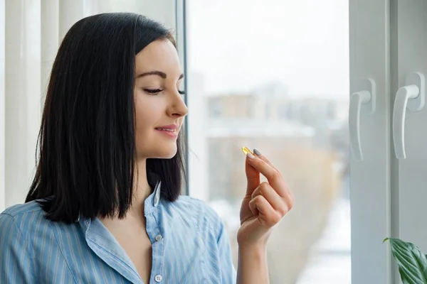 Junge Frau, die zu Hause am Fenster steht und Vitamin-Omega-3-Kapsel Fischöl einnimmt — Stockfoto